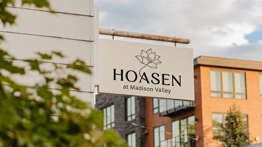 Hoa Sen at Madison Valley storefront sign mounted on a building exterior, with leafy greenery in the foreground and modern brick buildings visible in the background.