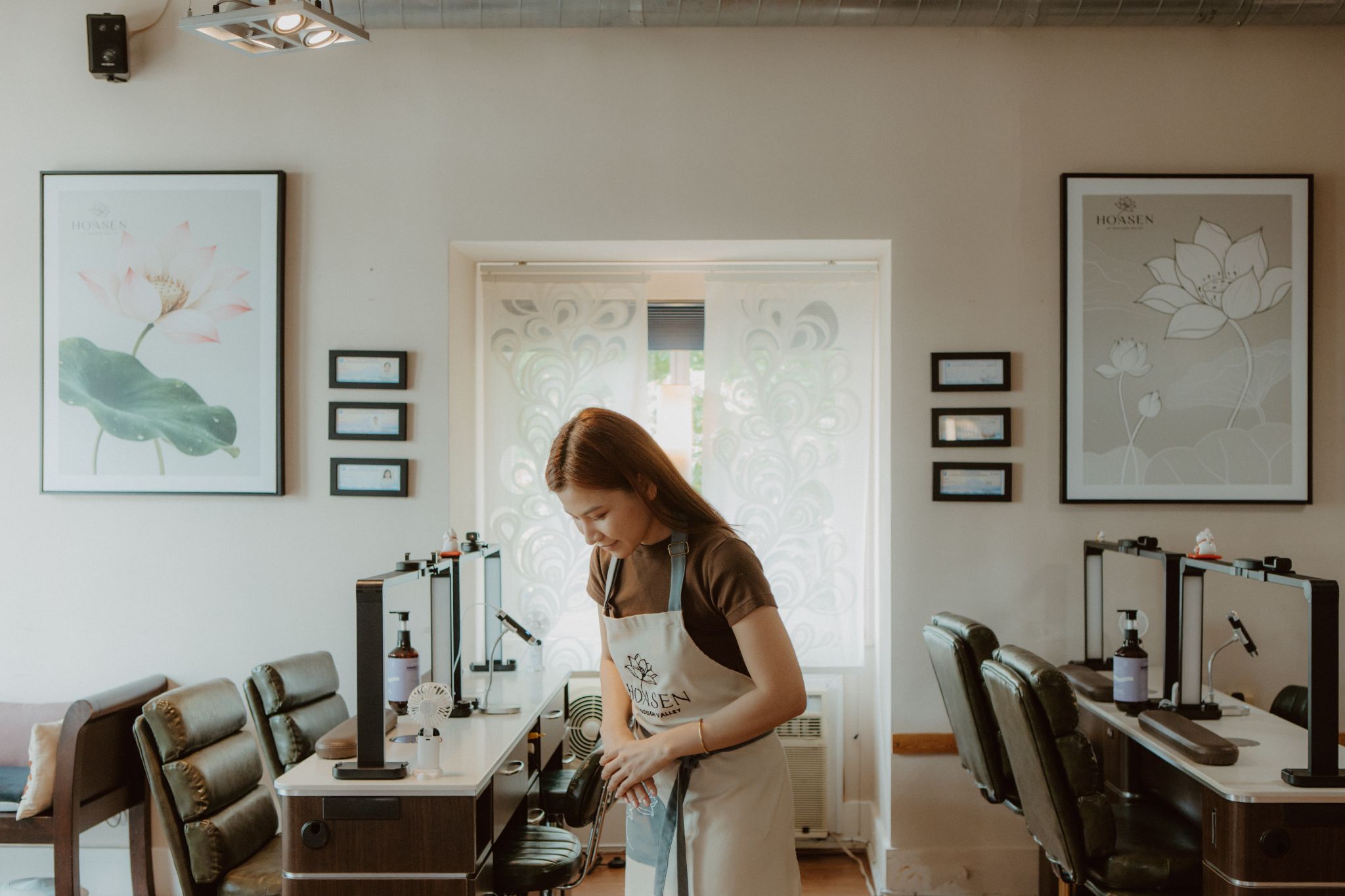 A nail technician wearing a Hoasen apron prepares a manicure station inside a bright, elegant nail salon, surrounded by leather chairs, manicure tables, framed lotus artwork, and soft natural lighting.