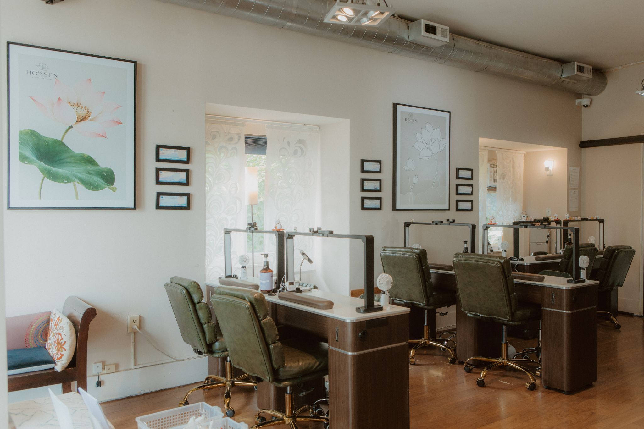 A bright, elegant nail salon interior with several manicure stations, dark green chairs, wooden tables, and glass partitions. Large lotus flower artwork decorates the white walls, while natural light enters through patterned windows, creating a calm and welcoming atmosphere.