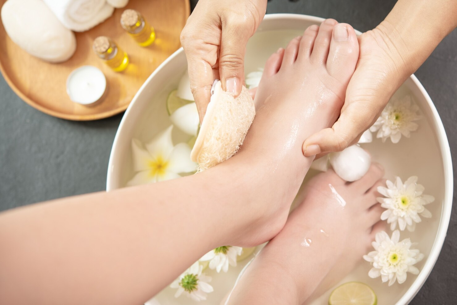 Hands exfoliating a foot during a spa pedicure treatment in a bowl of water with white flowers and spa accessories.