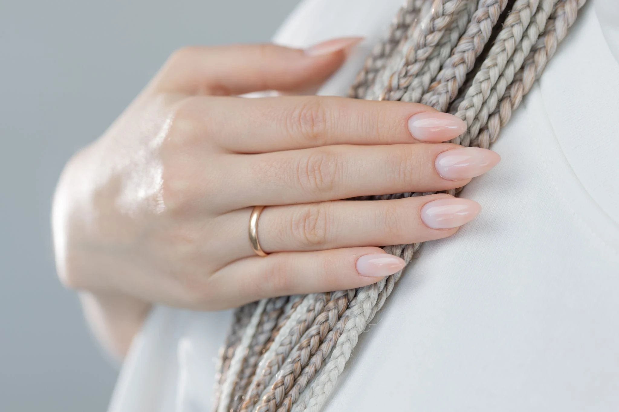 Hand with glossy nude pink almond-shaped nails resting on a light fabric surface, with braided rope detail in the background.