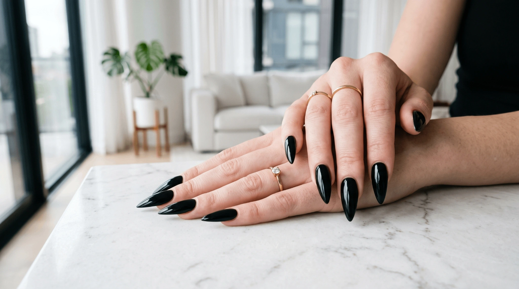 Hands with glossy black stiletto nails placed on a white marble surface near a bright window.