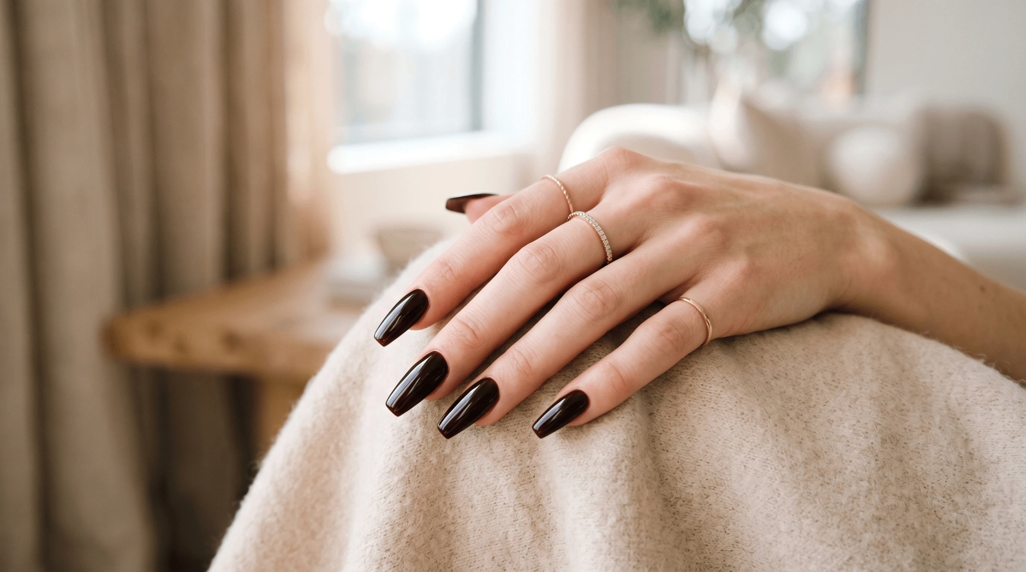 Hand with glossy dark brown ballerina-shaped nails resting on a light-colored blanket in a softly lit indoor setting.