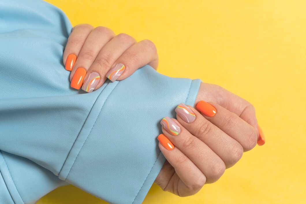 Hands with glossy orange and nude nail enhancements featuring abstract nail art, resting on a light blue sleeve against a yellow background.
